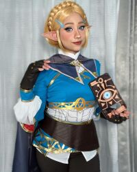 Rule 34 | 1girl, blonde hair, blue eyes, cosplay, female focus, fingerless gloves, gloves, highres, indoors, light smile, mexican (nationality), nintendo, photo (medium), pointy ears, princess zelda, short hair, solo, standing, taliverse, the legend of zelda, the legend of zelda: breath of the wild