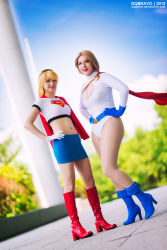 Rule 34 | 2girls, blonde hair, boots, cape, cosplay, dc comics, gloves, hands on own hips, highres, jenni hashimoto, leotard, multiple girls, photo (medium), power girl, s shield, smile, supergirl, superman (series)
