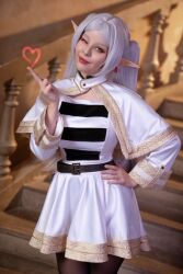 Rule 34 | 1girl, cosplay, dress, earrings, elf, female focus, frieren, frieren (cosplay), heart, helly von valentine (disharmonica), jewelry, looking at viewer, one eye closed, photo (medium), pointy ears, real life, red lips, skirt, solo, sousou no frieren, white hair, wink