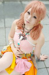 Rule 34 | 1girl, belt, bracelet, cosplay, female focus, final fantasy, final fantasy xiii, jewelry, looking at viewer, midriff, necklace, oerba dia vanille, oerba dia vanille (cosplay), on floor, outdoors, pink shirt, shirt, sitting, skirt, solo, tiles, twintails