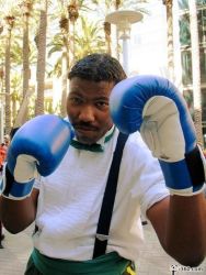 Rule 34 | 1boy, blouse, bow, bowtie, boxing gloves, capcom, cosplay, cosplay photo, dudley, dudley (cosplay), gloves, green bow, green bowtie, male focus, palm tree, photo (medium), shirt, solo, street fighter, street fighter iii (series), tree, watermark, web address, white shirt