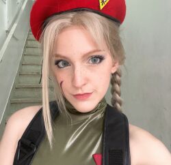 Rule 34 | 1girl, :&gt;, bare shoulders, blonde hair, blue eyes, braid, brazilian (nationality), breasts, cammy white, cammy white (cosplay), cosplay, freckles, green leotard, hat, highres, leotard, long hair, memepantazis, pale skin, real life, sleeveless, smile, street fighter, street fighter 6, twin braids, twitter username, upper body